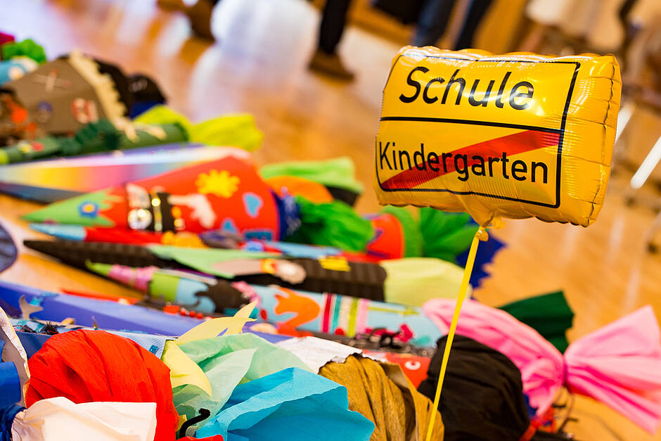 Man sieht auf Schultüten der Schulneulinge, zwischen denen ein Luftballon herausragt, auf dem - wie bei einem Ortsendeschild - das Wort KINDERGARTEN durchgestrichen ist und darüber das Wort SCHULE zu erkennen ist.